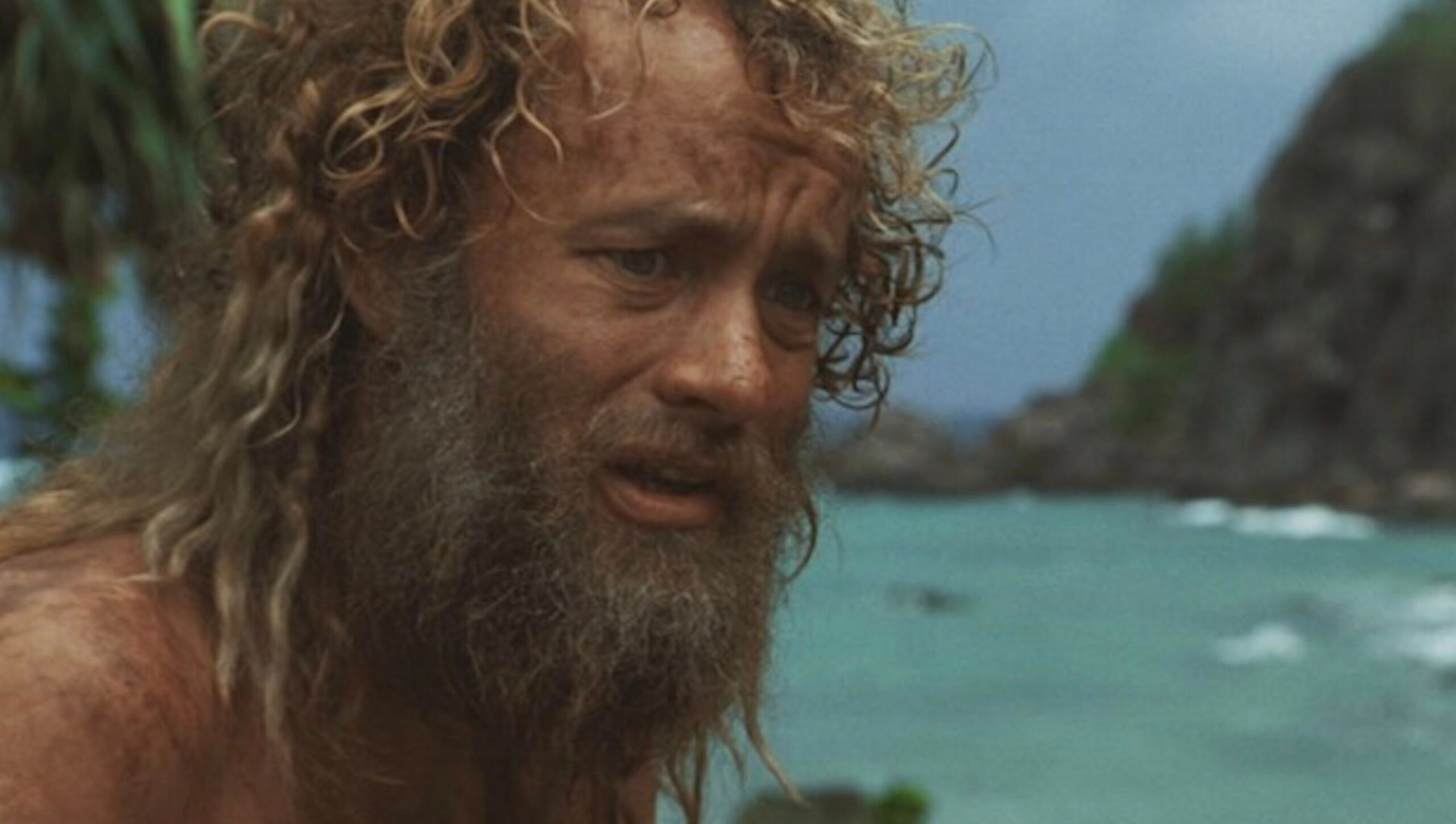 A man with long curly hair and a thick beard looks distressed while standing by the ocean, with lush greenery and rocky cliffs in the background.