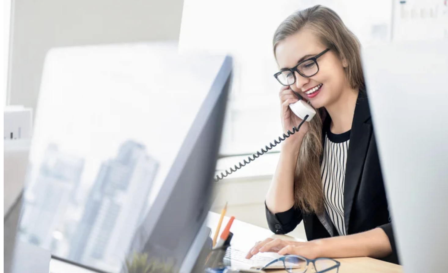 A smiling woman wearing glasses and a black blazer talks on a landline phone at her desk in a bright office, with computer monitors and city buildings visible in the background.