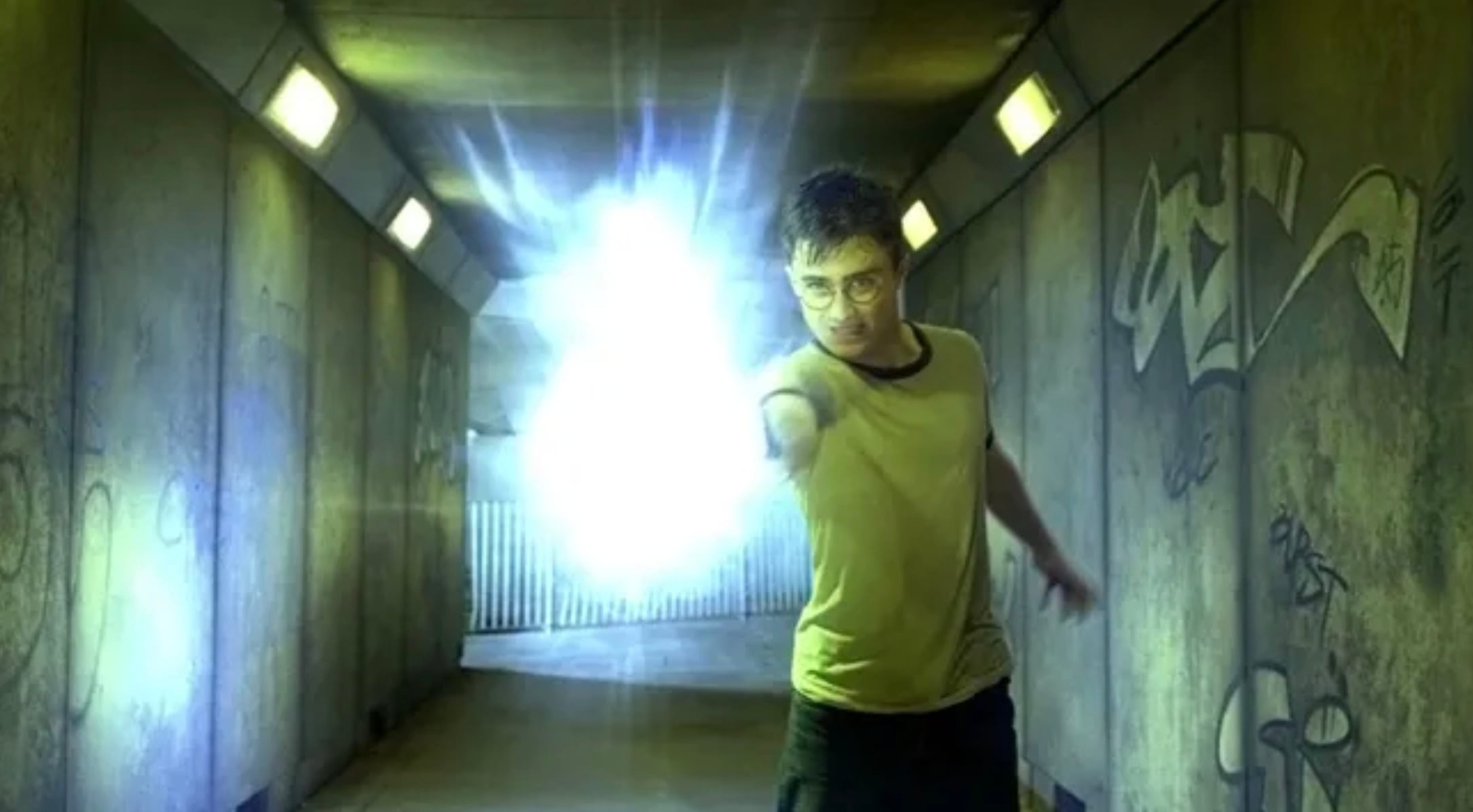 A young man in glasses stands in a graffiti-covered tunnel, extending his arm forward as he casts a bright, glowing blue spell or energy from his hand. The light illuminates the tunnel around him.