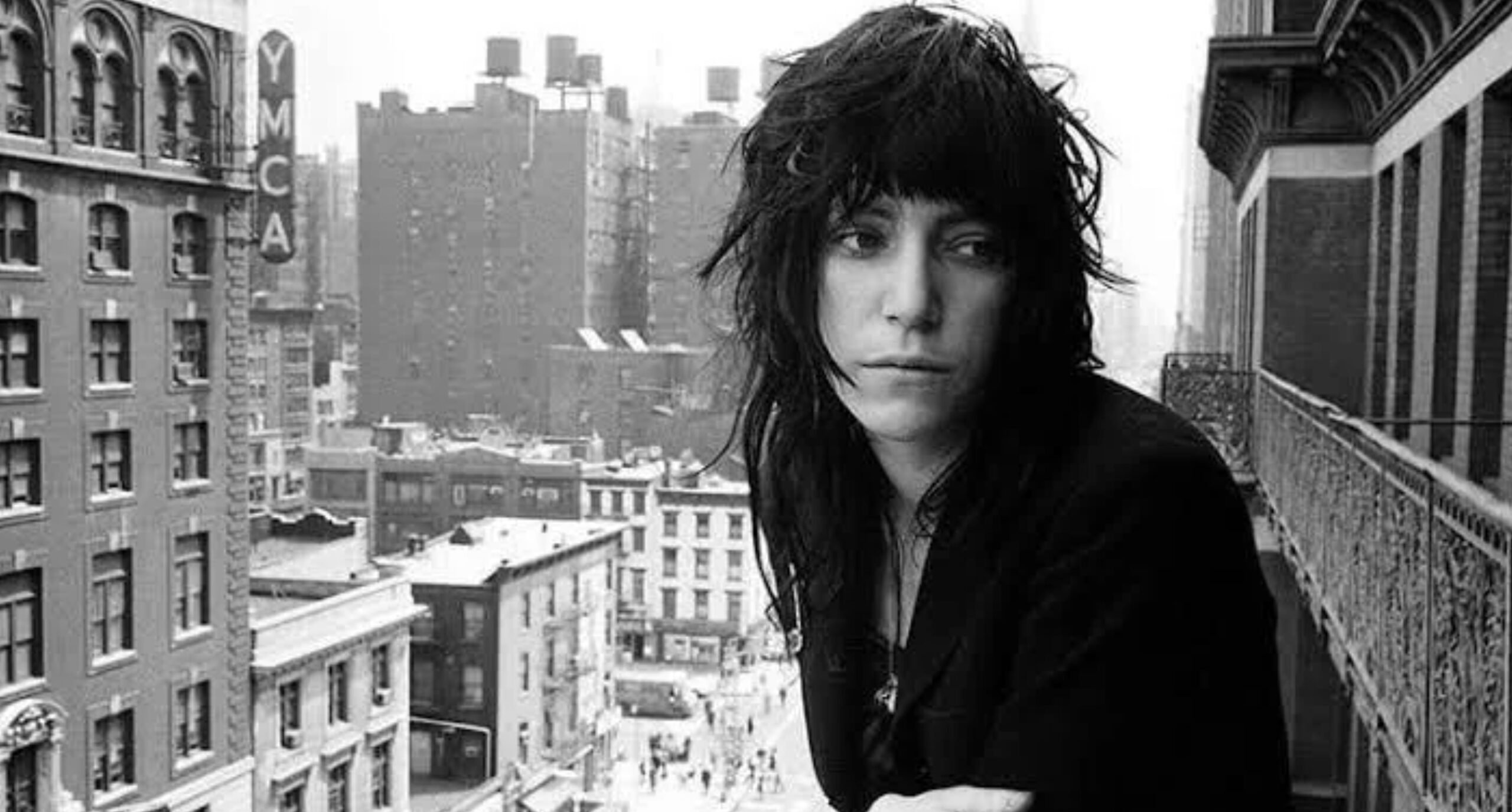 A person with long, tousled dark hair and a serious expression leans on a balcony railing overlooking a city street lined with tall buildings. The photo is black and white, capturing an urban scene.