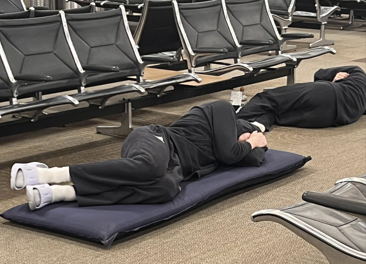 Two people in dark clothing are lying on mats on the floor of an airport terminal, using their arms to cover their faces, with empty rows of black chairs beside them.