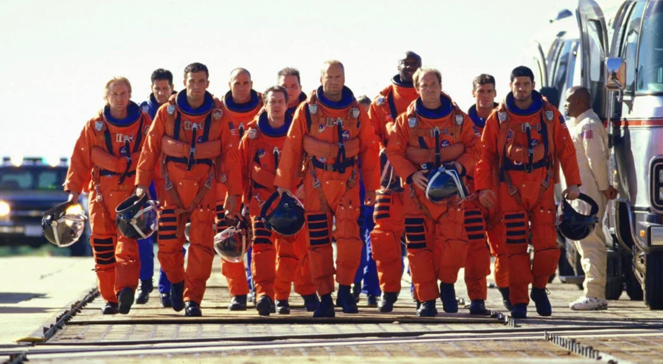 A group of astronauts in orange space suits walk side by side outdoors, holding their helmets and heading toward a bus, with clear skies in the background.