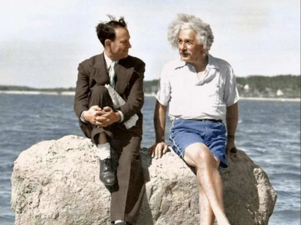 Two men sit on a large rock by the water; one wears a suit and tie, the other a white shirt and blue shorts. They appear to be talking, with a scenic landscape and trees visible in the background.