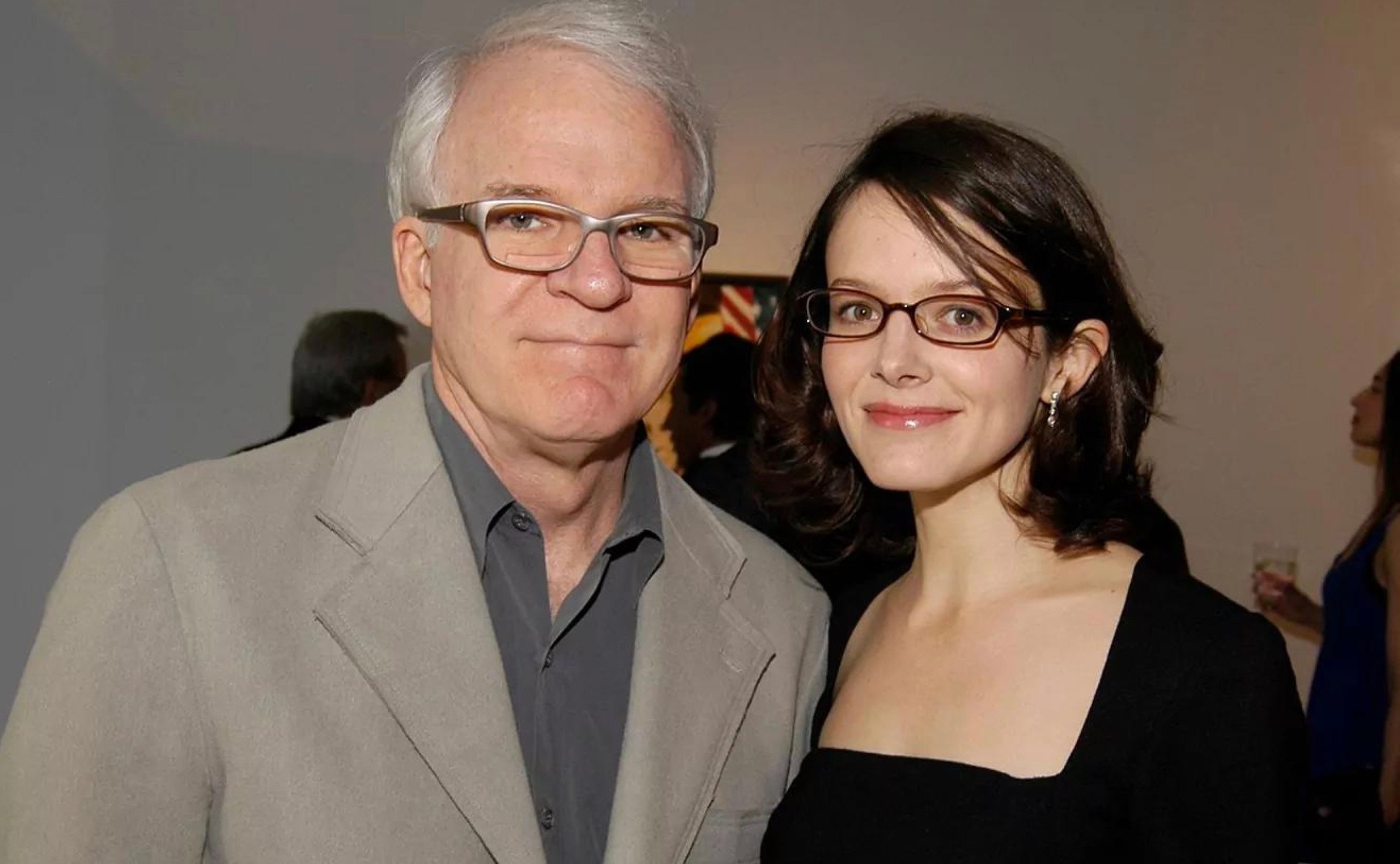 Two people are standing together and smiling at the camera at an indoor event. The man has gray hair, glasses, and is wearing a gray suit, while the woman has brown hair, glasses, and is wearing a black dress.