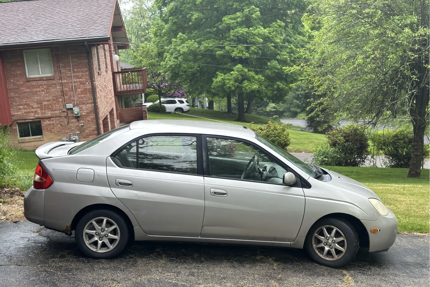 A silver sedan is parked on a driveway in front of a brick house, with green trees and grass visible in the background on a cloudy day.