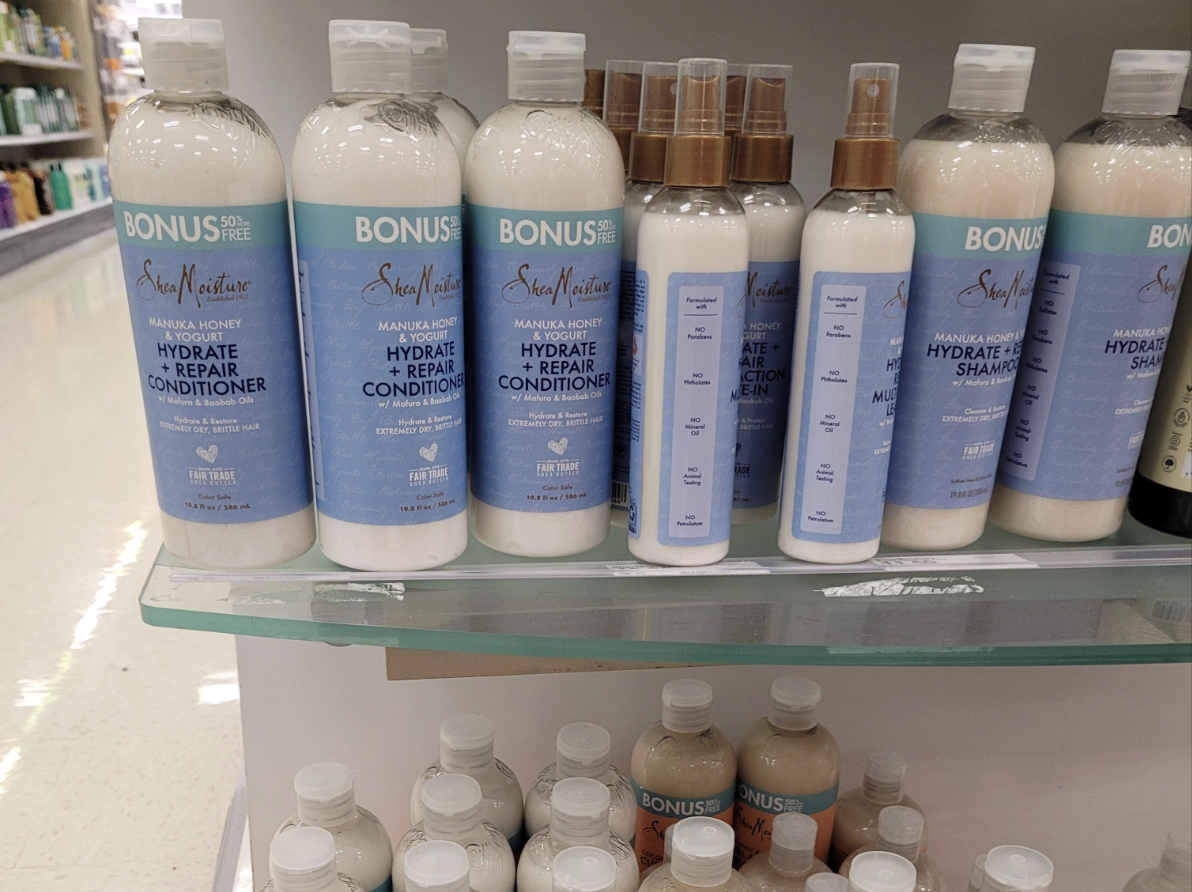 Several bottles of SheaMoisture Manuka Honey Hydrate + Repair Conditioner and matching spray mist are displayed on a store shelf, with bonus size labels. Smaller bottles are seen on the lower shelf.