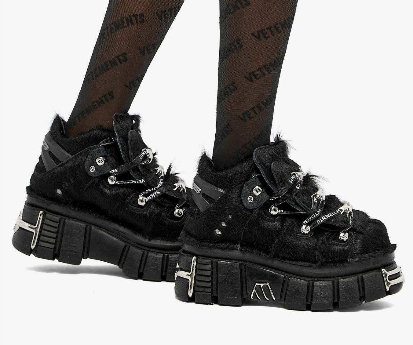 A person wearing black platform sneakers with chunky soles, faux fur details, and silver hardware, paired with black tights featuring the "VETEMENTS" logo in a repeating pattern.