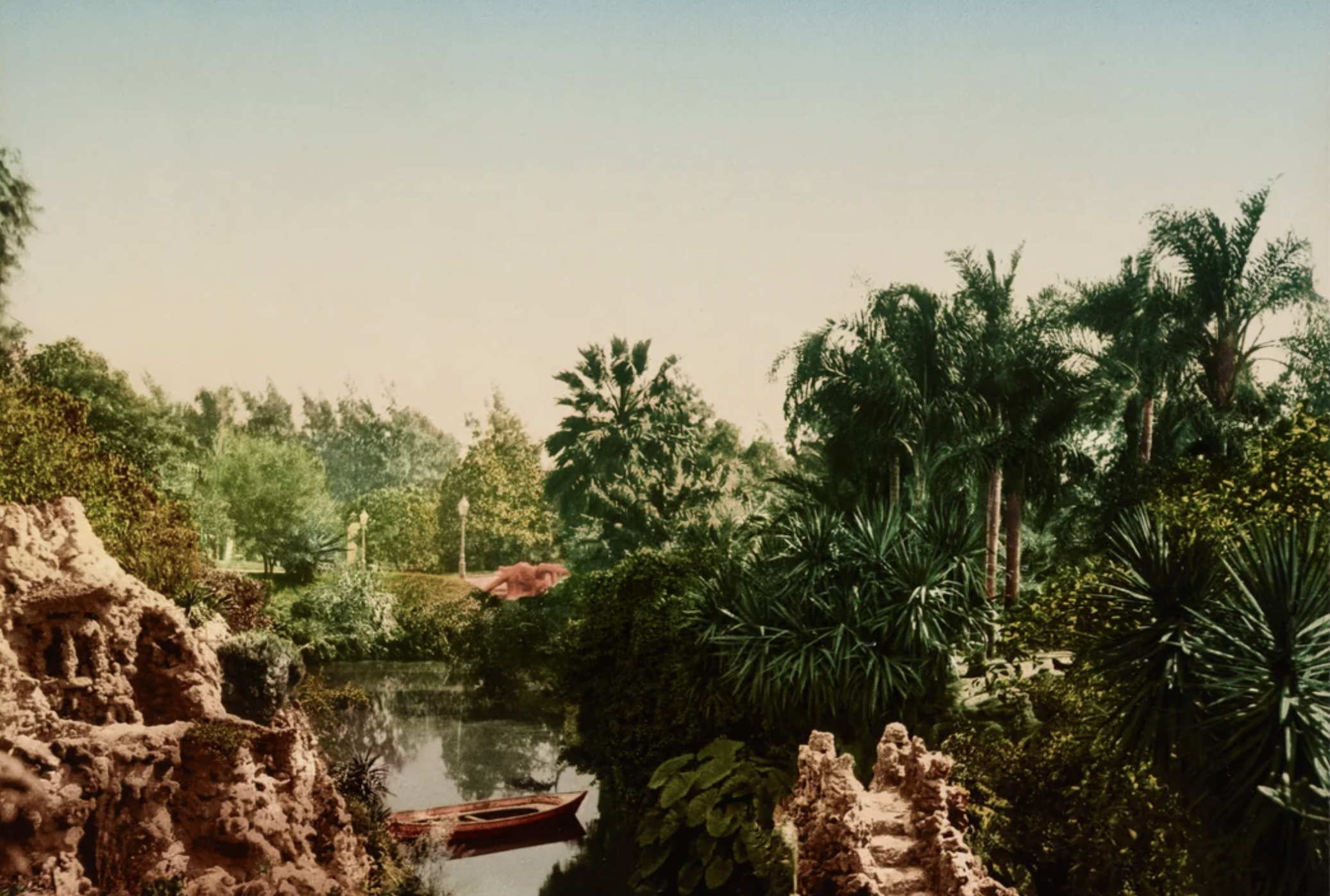 A lush tropical garden with palm trees and dense greenery surrounds a small pond. A red rowboat floats on the water, and rocky formations frame the scene under a clear sky.