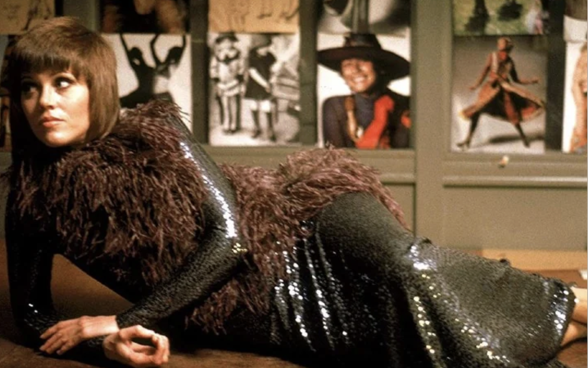 A woman with short brown hair reclines on her side, wearing a sparkly black gown with textured fur-like details. Behind her, a wall displays various fashion sketches and photographs.