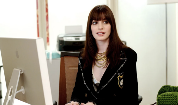 A young woman with long brown hair and bangs sits at an office desk, wearing a black blazer with a crest, layered necklaces, and a white top, looking to the side with a slight, uncertain smile.