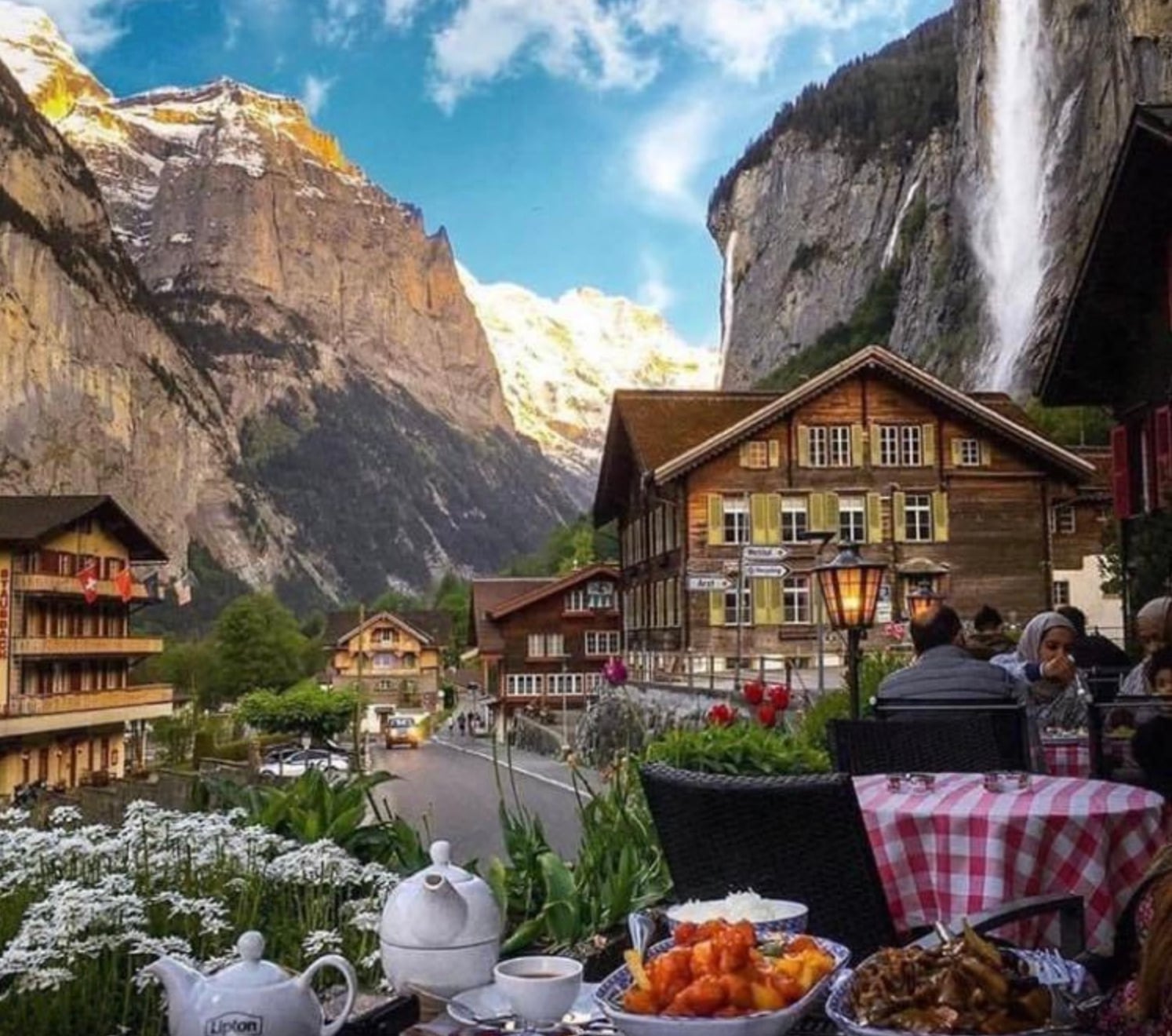A cozy outdoor café with red checkered tablecloths overlooks a scenic mountain village, wooden chalets, a tall waterfall, and snow-capped peaks under a blue sky. Plates of food and tea are set on the table.