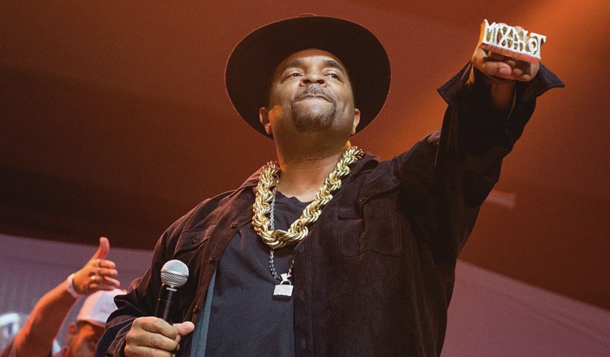 A man wearing a black hat, gold chain, and dark clothing holds a microphone in one hand and raises the other, showing a large ring. The background is lit with orange and pink stage lights.