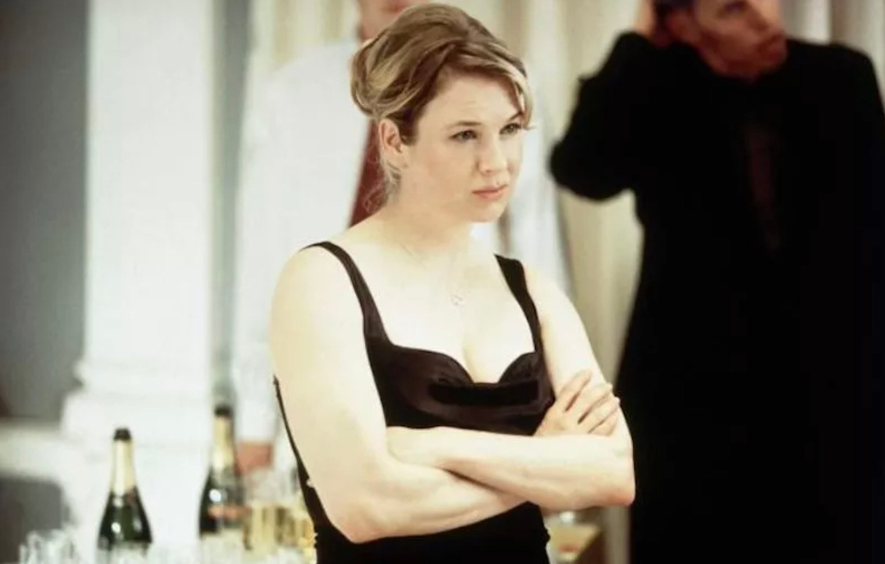 A woman in a black dress stands with her arms crossed, looking slightly to the side with a neutral expression. Bottles and glasses are on the table beside her, and blurred people are visible in the background.