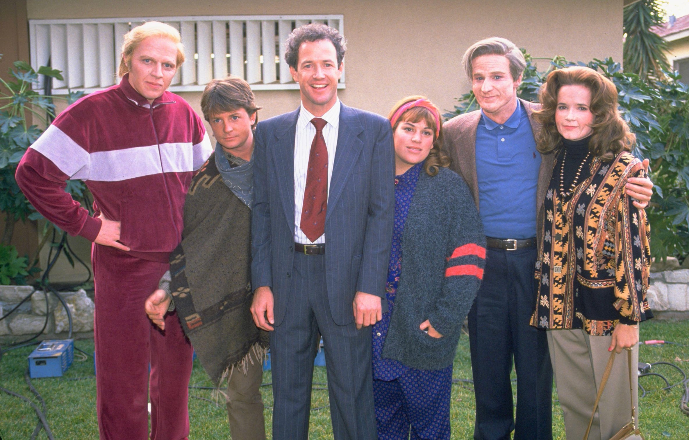 Six people stand together outside in a yard, posing for a photo. They are dressed in various costumes, some colorful and retro, and all are smiling or looking at the camera, with greenery and a house in the background.