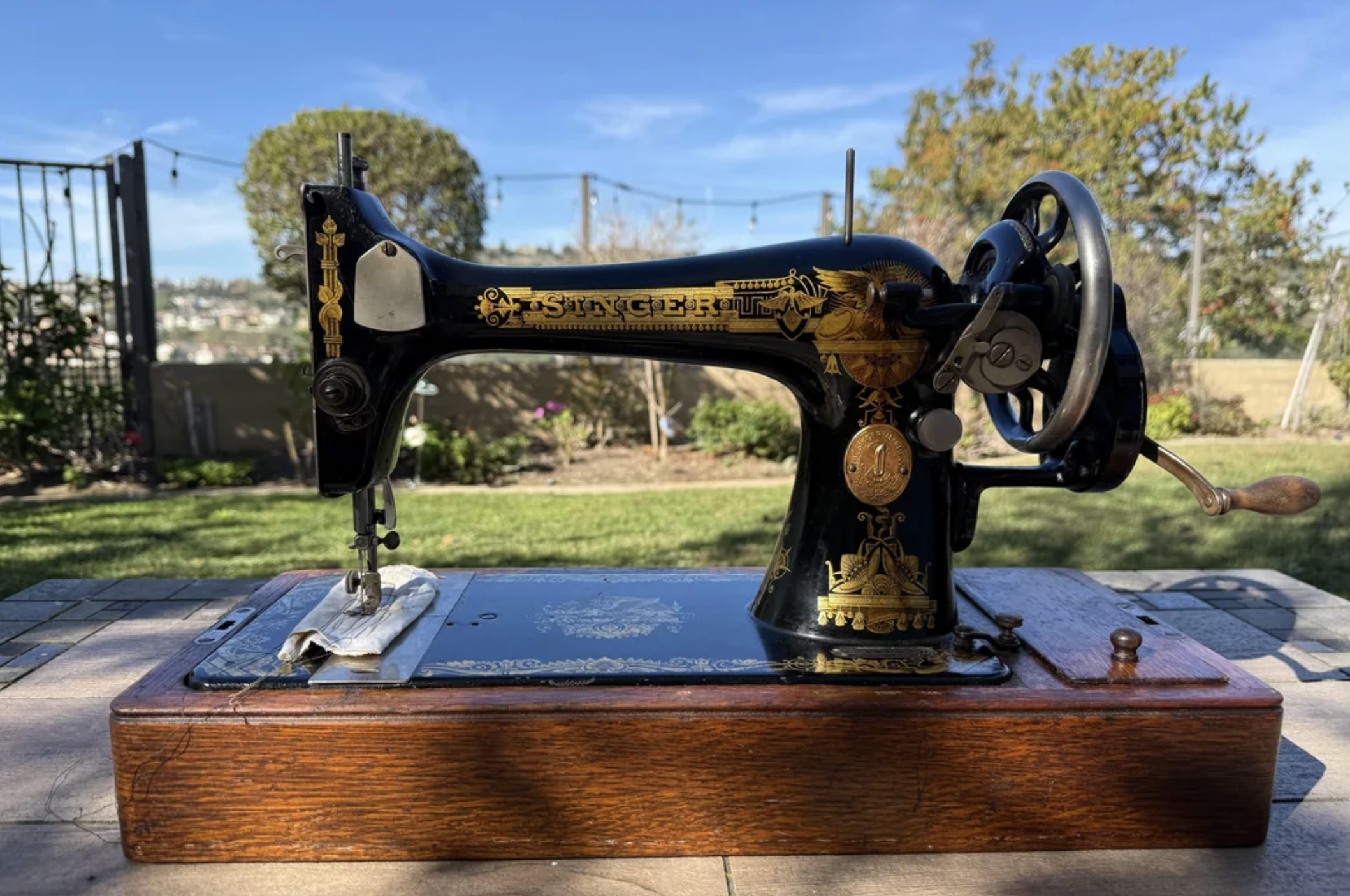 A vintage black and gold Singer sewing machine sits on a wooden base outdoors, with a garden and fence in the background under a sunny sky.