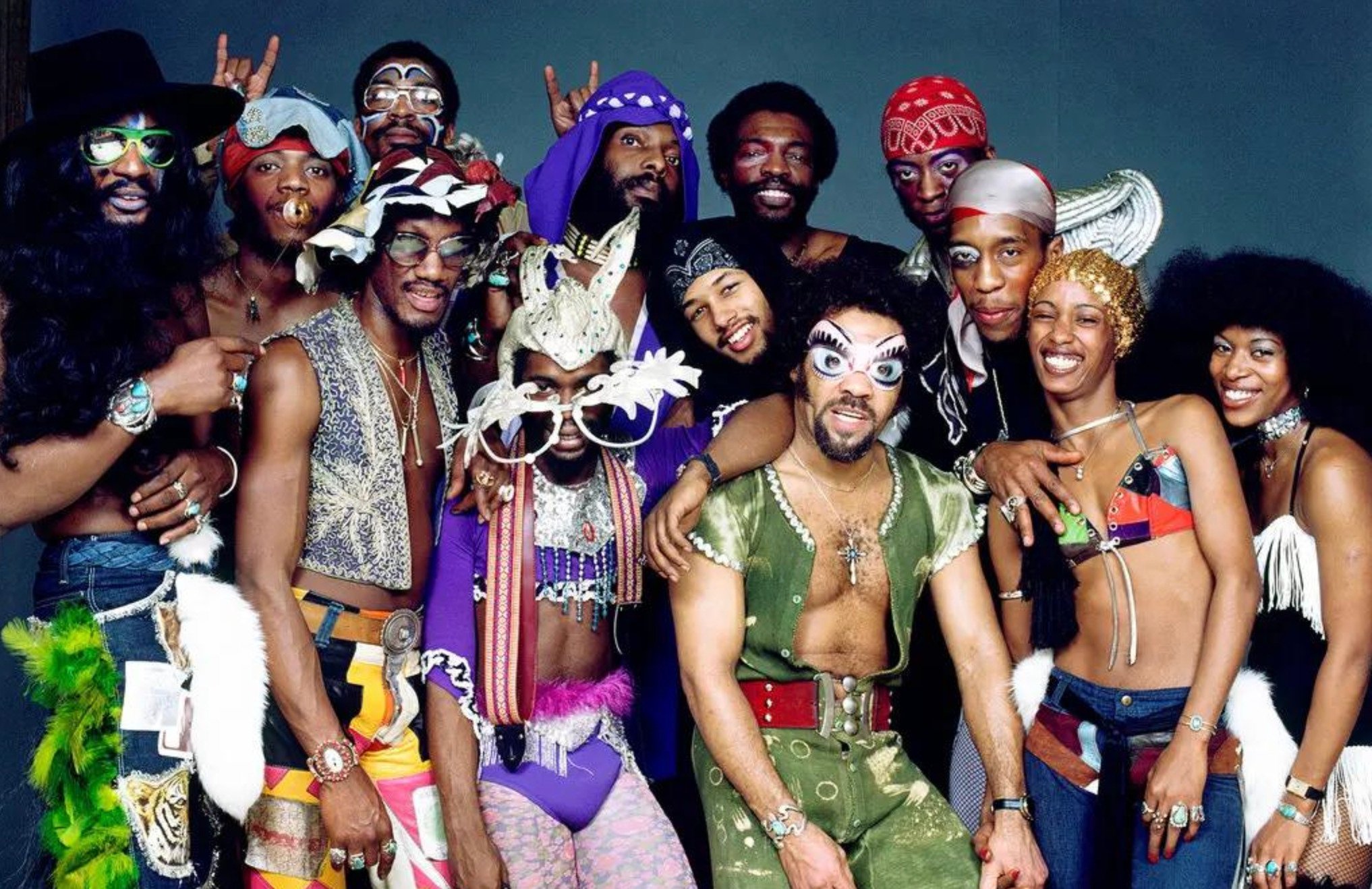 A group of people pose together wearing colorful, eclectic, and funky costumes with vibrant patterns, feathers, headbands, sunglasses, hats, and bold accessories, evoking a playful 1970s funk or disco style.
