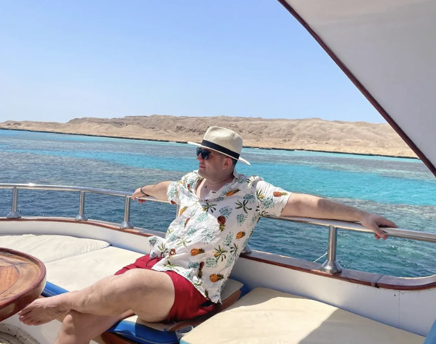 A man in a hat, sunglasses, a tropical shirt, and red shorts relaxes on a boat with turquoise water and rocky, sandy land visible in the background under a clear blue sky.