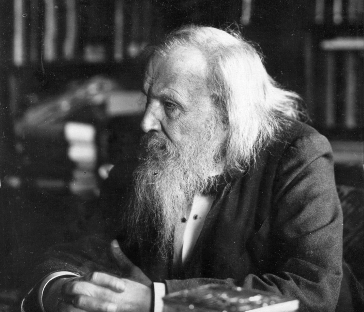An older man with long white hair and a full beard sits indoors, wearing a dark suit. He is in profile, facing left, with his hands clasped. Books and shelves are visible in the background.