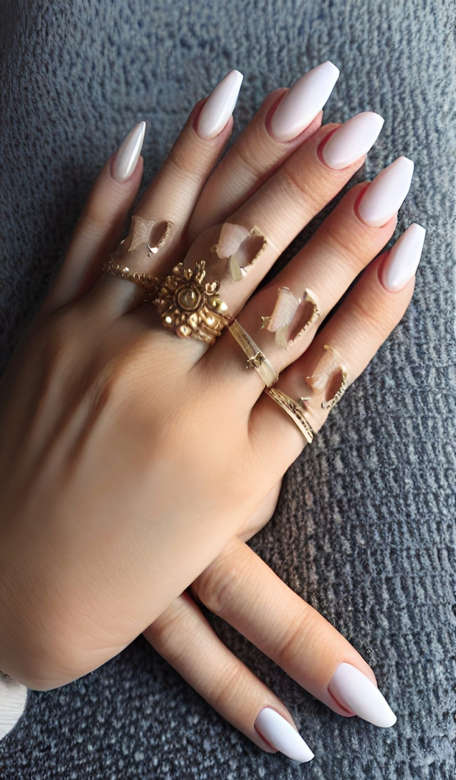 A close-up of hands with long, almond-shaped, light pink nails featuring subtle white tips. The fingers are adorned with multiple gold rings, and the background is a textured gray fabric.