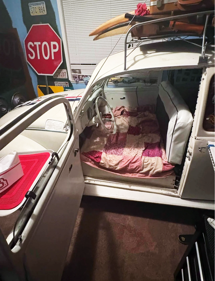 A white vintage car with its door open is parked indoors; the backseat and floor are covered with a pink patchwork quilt, and surfboards are strapped to the roof. The room has a STOP sign and various decorations on the walls.