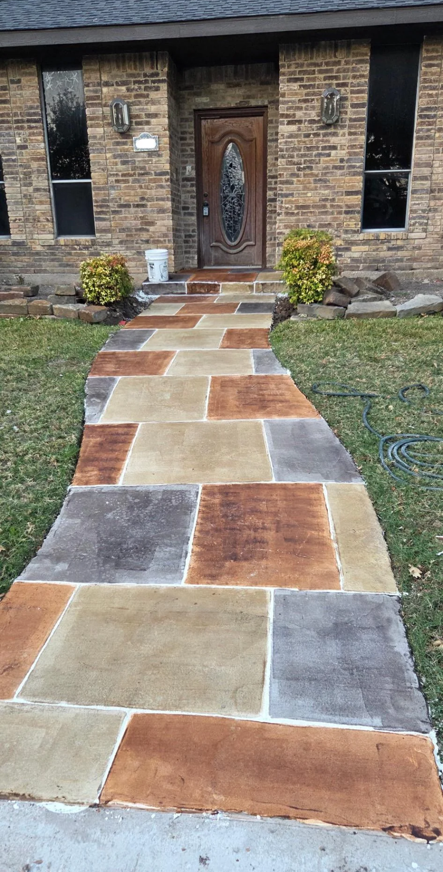 A curved walkway made of large, rectangular tiles in brown, tan, and gray shades leads to a wooden front door of a brick house, with small shrubs on either side and grass along the path.