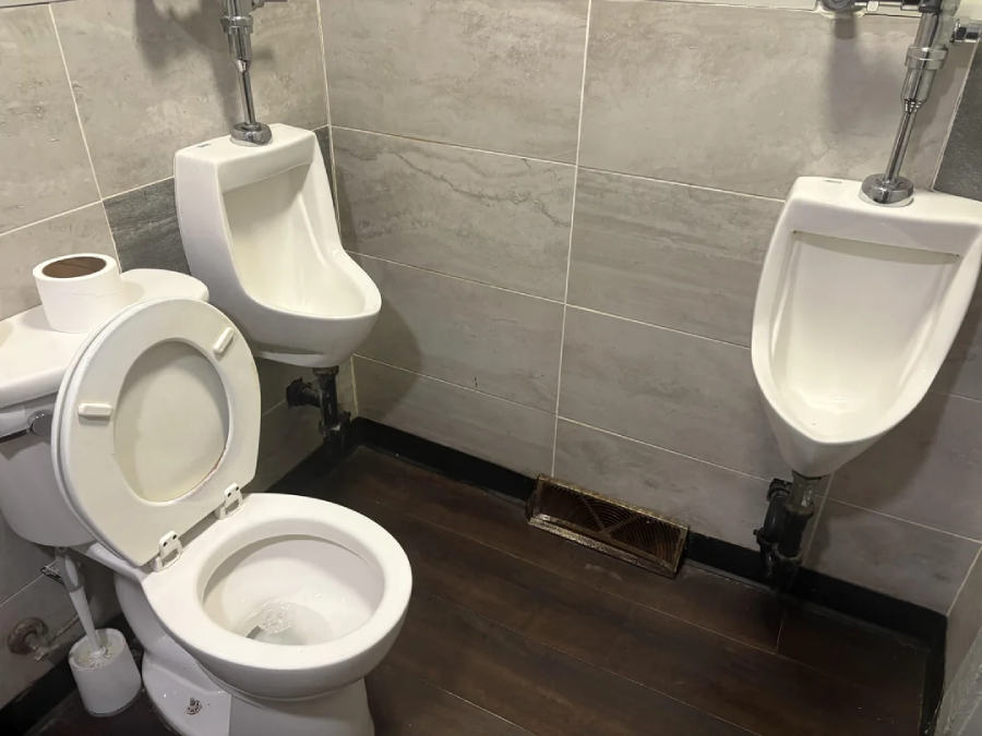 A restroom with two urinals and one toilet positioned close together in a corner, with a roll of toilet paper placed on the toilet tank. The walls are tiled and the floor is dark wood.