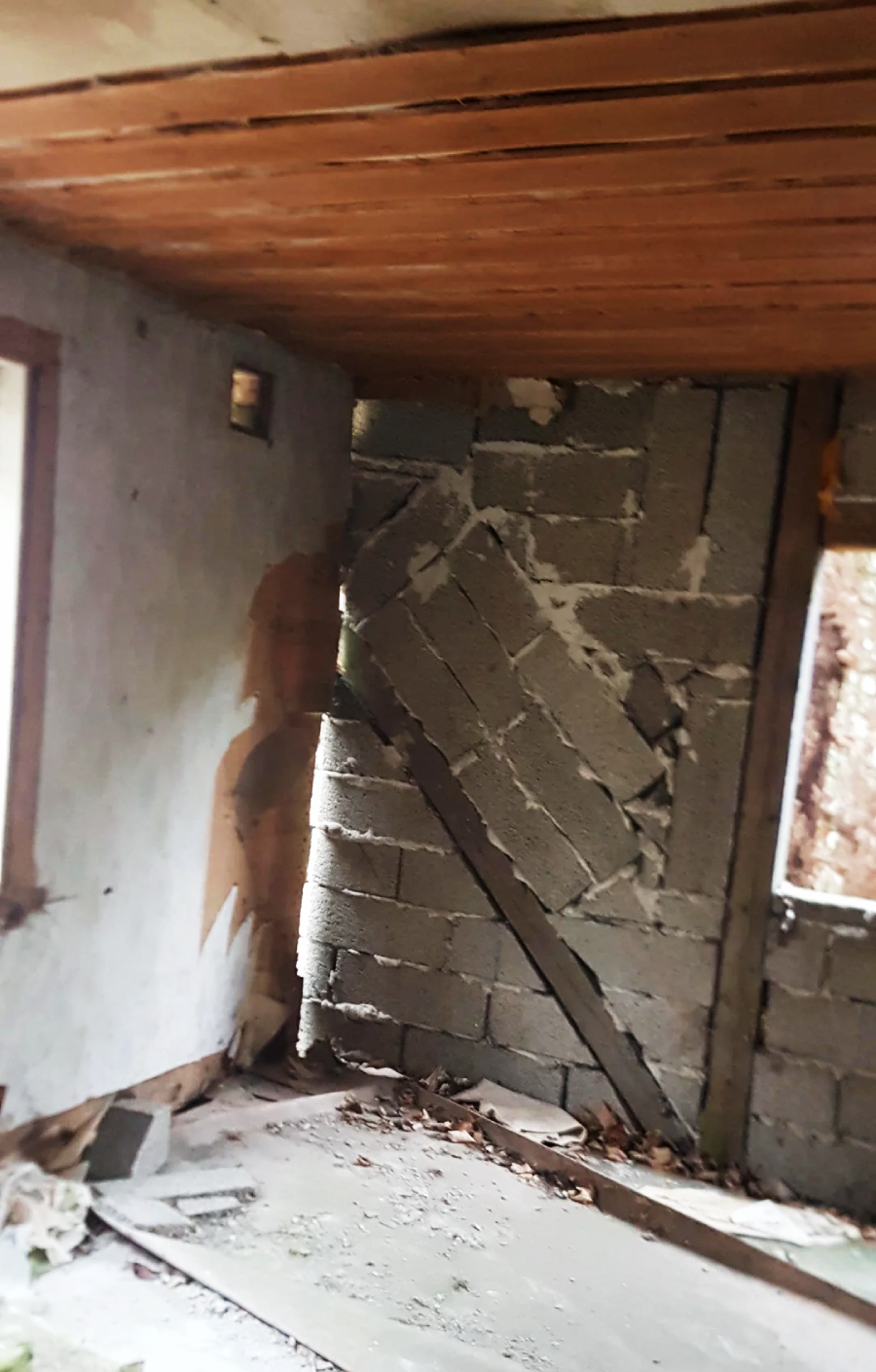 A damaged interior wall of a building, made of cinder blocks, partially collapsed and tilted. The room has a wooden ceiling, dirty floor, and two windows allowing light inside. The overall space looks abandoned and in disrepair.