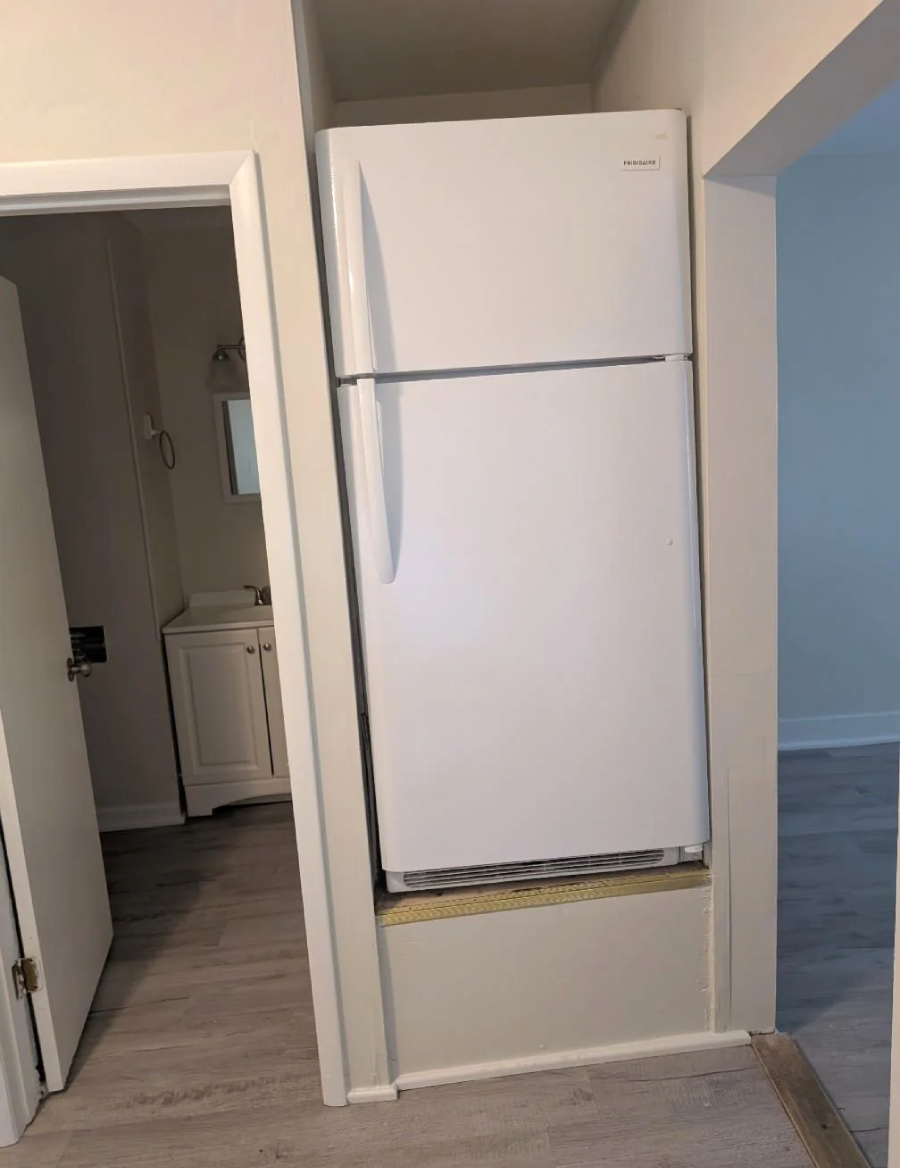 A white refrigerator awkwardly installed in a recessed wall space, raised off the ground with a visible gap beneath. The fridge is next to a doorway leading to a small bathroom with a sink and mirror.