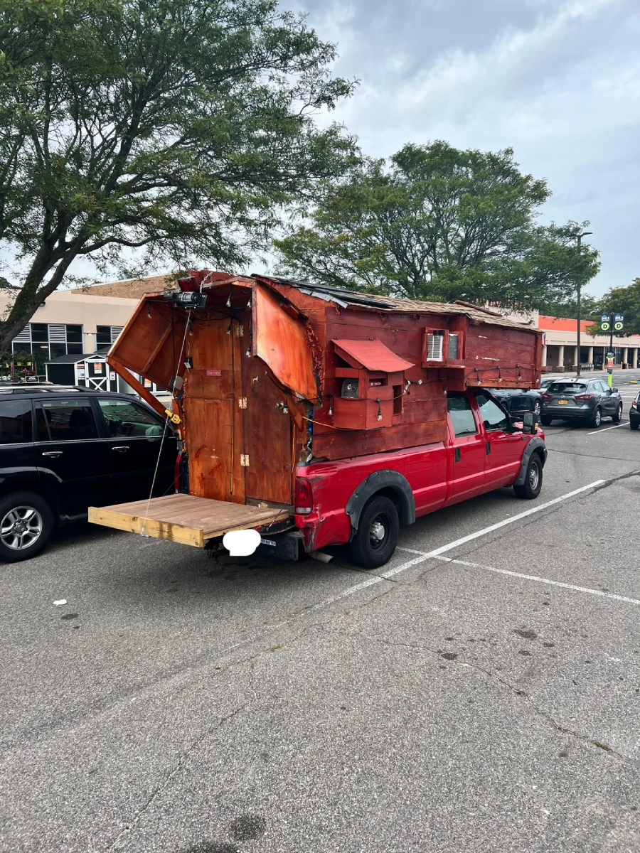 A red pickup truck with a large, homemade wooden cabin structure built onto its bed, featuring windows, a door, and a plywood platform extending from the back, is parked in a lot among other cars.