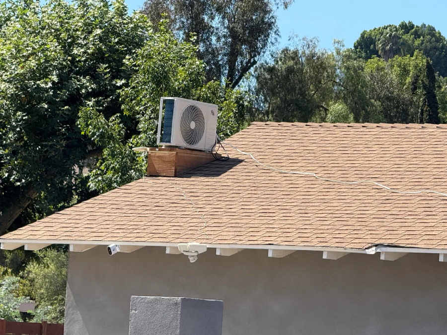 A rooftop air conditioning unit is mounted on a small platform on a house with brown shingles. Wires run across the roof, and trees are visible in the background under a clear blue sky.