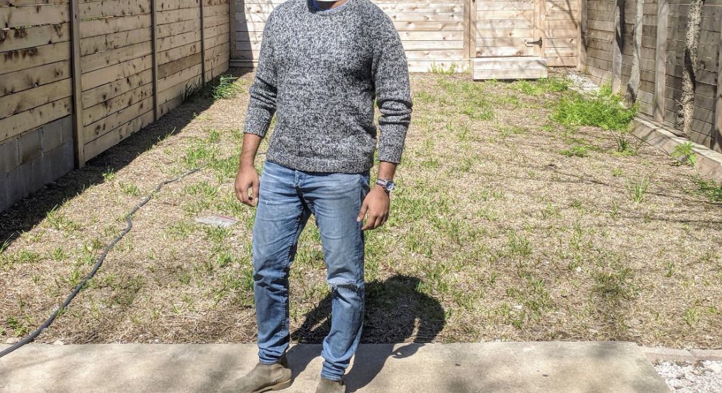 A person stands on a concrete path in a sunlit backyard with patchy grass, wearing a gray sweater, blue jeans, brown shoes, and a watch. Wooden fences line the yard, and shadows fall across the scene.