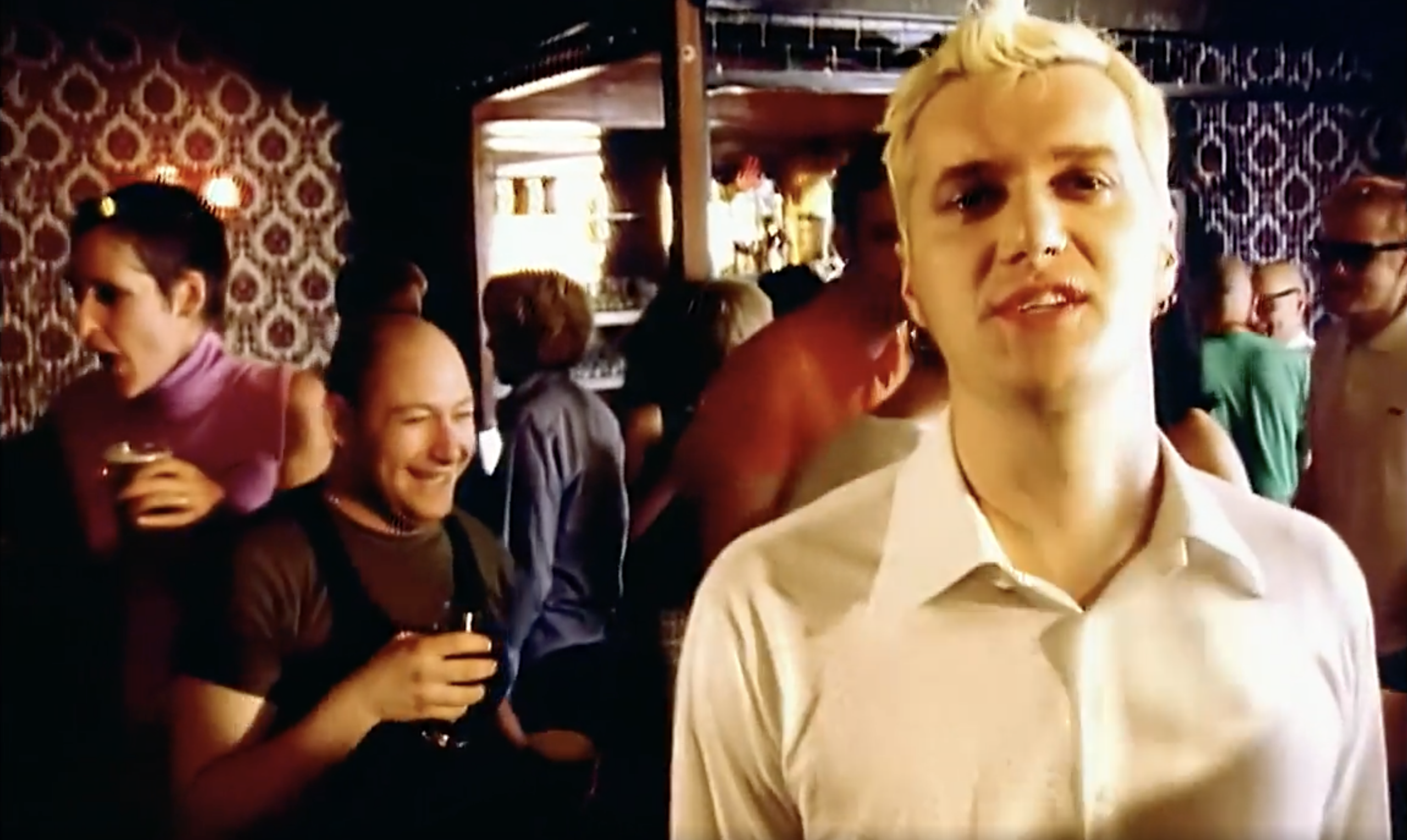 A man with short blond hair wearing a white shirt stands in the foreground of a crowded bar with patterned wallpaper; several people are socializing and holding drinks in the background.