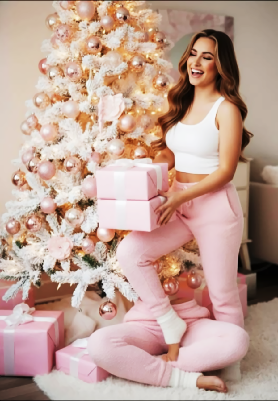 A smiling woman in a white crop top and pink sweatpants stands beside a white Christmas tree decorated with pink ornaments, holding pink gift boxes. Wrapped presents are arranged under the tree.