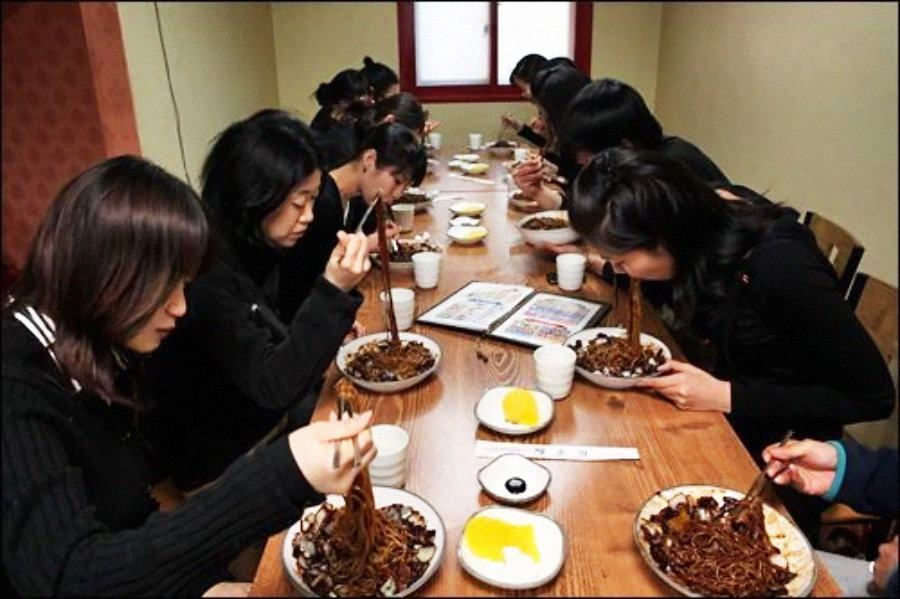 A group of people sit at a long wooden table in a restaurant, eating noodles with chopsticks. Plates with yellow pickled radish and cups are on the table. Most diners have dark hair and are focused on their food.