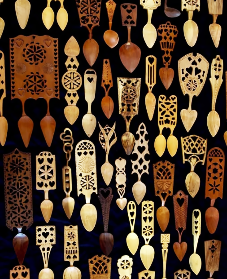 A collection of intricately carved wooden spoons and spatulas, featuring various shapes, patterns, and heart motifs, arranged vertically against a dark background.