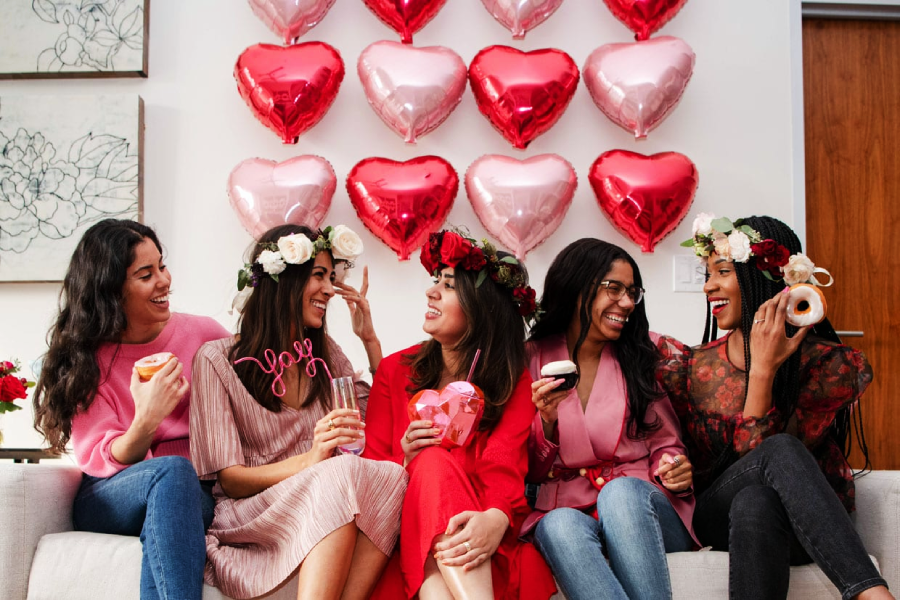 Five women sit on a couch smiling and laughing, holding donuts, drinks, and heart-shaped props. They wear flower crowns and are in front of a wall decorated with pink and red heart balloons.