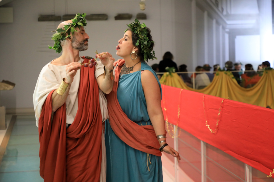 Two people dressed in ancient Roman-style costumes with leafy crowns stand indoors, holding grapes. One wears a red and white toga, the other a blue dress. Colorful drapes and a group of people are visible in the background.
