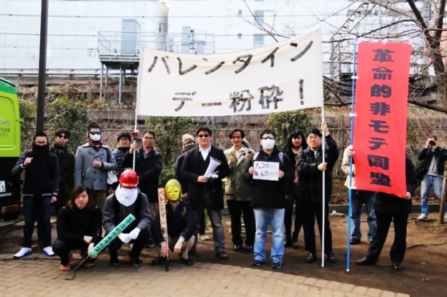 A group of people pose outdoors holding a large Japanese banner and various signs. Some wear masks, costumes, or sunglasses. The scene appears to be a protest or demonstration. Trees and an industrial building are in the background.