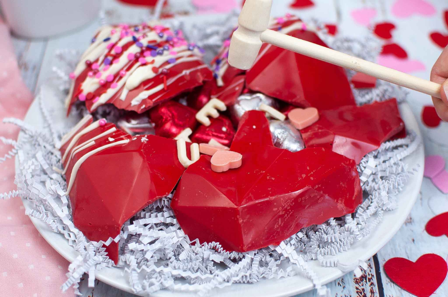 A plate with red geometric chocolate hearts, some broken open to reveal candy inside, surrounded by white shredded paper. A hand holds a small wooden mallet above the plate. Pink and red heart decorations are in the background.