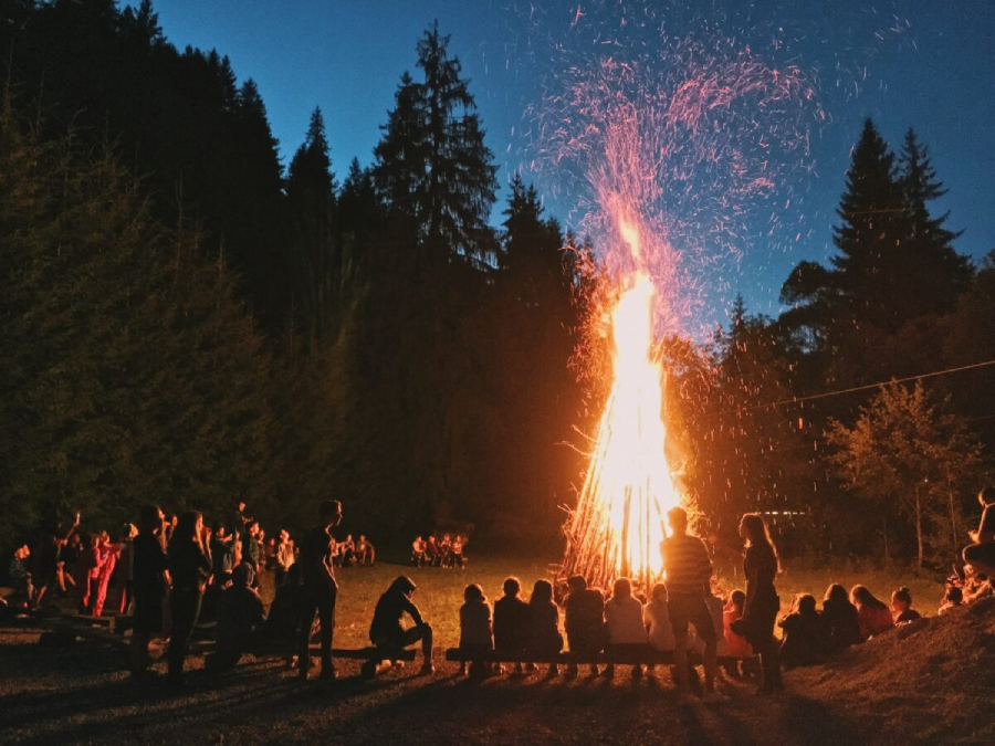 A group of people, including children, sit and stand around a large bonfire at dusk in a forest clearing. Sparks rise into the air as the sky darkens, and tall trees silhouette in the background.