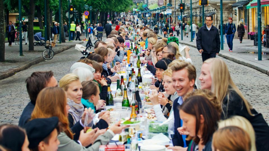 A large group of people sit together at a long outdoor table set with food and drinks, enjoying a communal meal on a city street lined with trees and buildings.