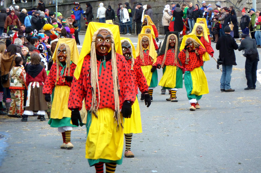 People in colorful costumes with yellow skirts, red and black spotted tops, yellow headscarves, striped socks, and large scary masks parade down a street, watched by a crowd of onlookers in the background.