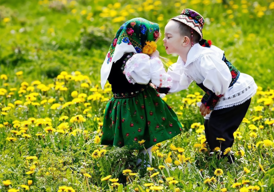 Two young children in traditional folk costumes stand in a field of yellow dandelions. The girl wears a green skirt and floral headscarf, while the boy leans in to kiss her, both smiling amid the flowers.