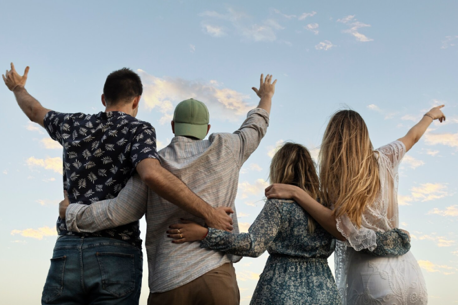 Four people stand side by side with their backs to the camera, arms around each other, raising their hands and pointing towards the sky under a clear, blue sky with some scattered clouds.