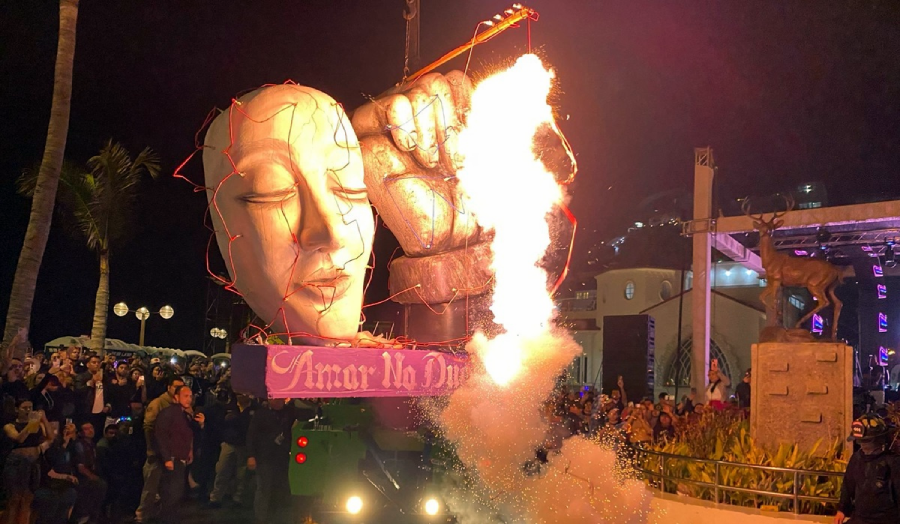 A large parade float featuring a serene human face and a raised fist emits flames at night. A crowd watches the spectacle, with a sign reading "Amar Na Duna" and city lights in the background.