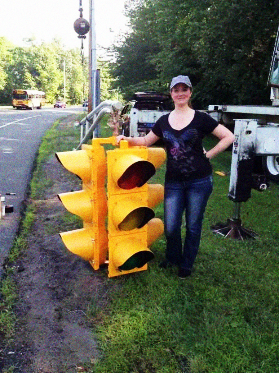 A woman in a cap stands outdoors beside a large, yellow traffic signal placed on grass near a roadside. There are trees, a school bus, and utility equipment visible in the background.
