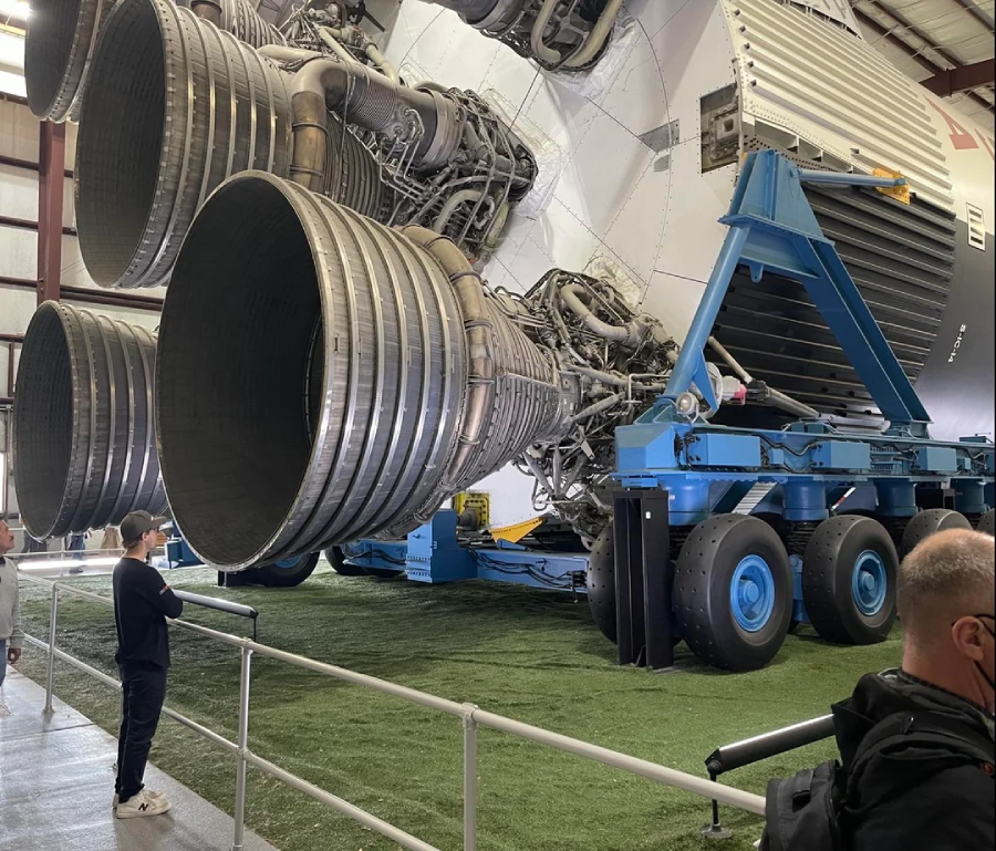 A large rocket engine on display indoors, with several people standing nearby for scale. The engine is mounted on a blue transport platform, and the area is fenced off with artificial grass beneath it.