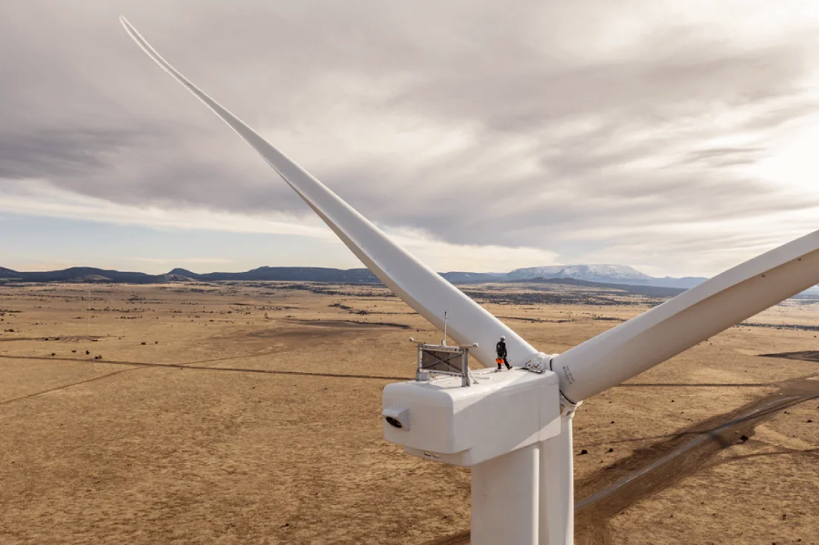 A person stands on top of a large white wind turbine, surrounded by a vast, dry landscape with mountains in the distance under a cloudy sky.