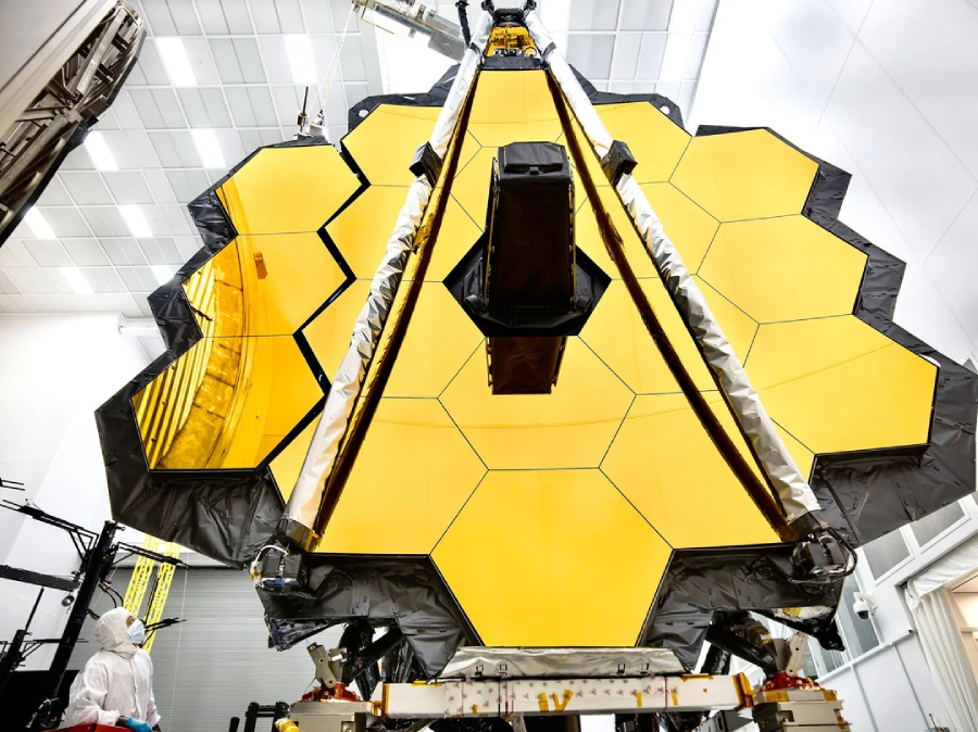 A large, gold hexagonal mirror structure of a space telescope is being assembled in a clean room. A person in a white protective suit stands nearby for scale. The surroundings are bright and sterile.