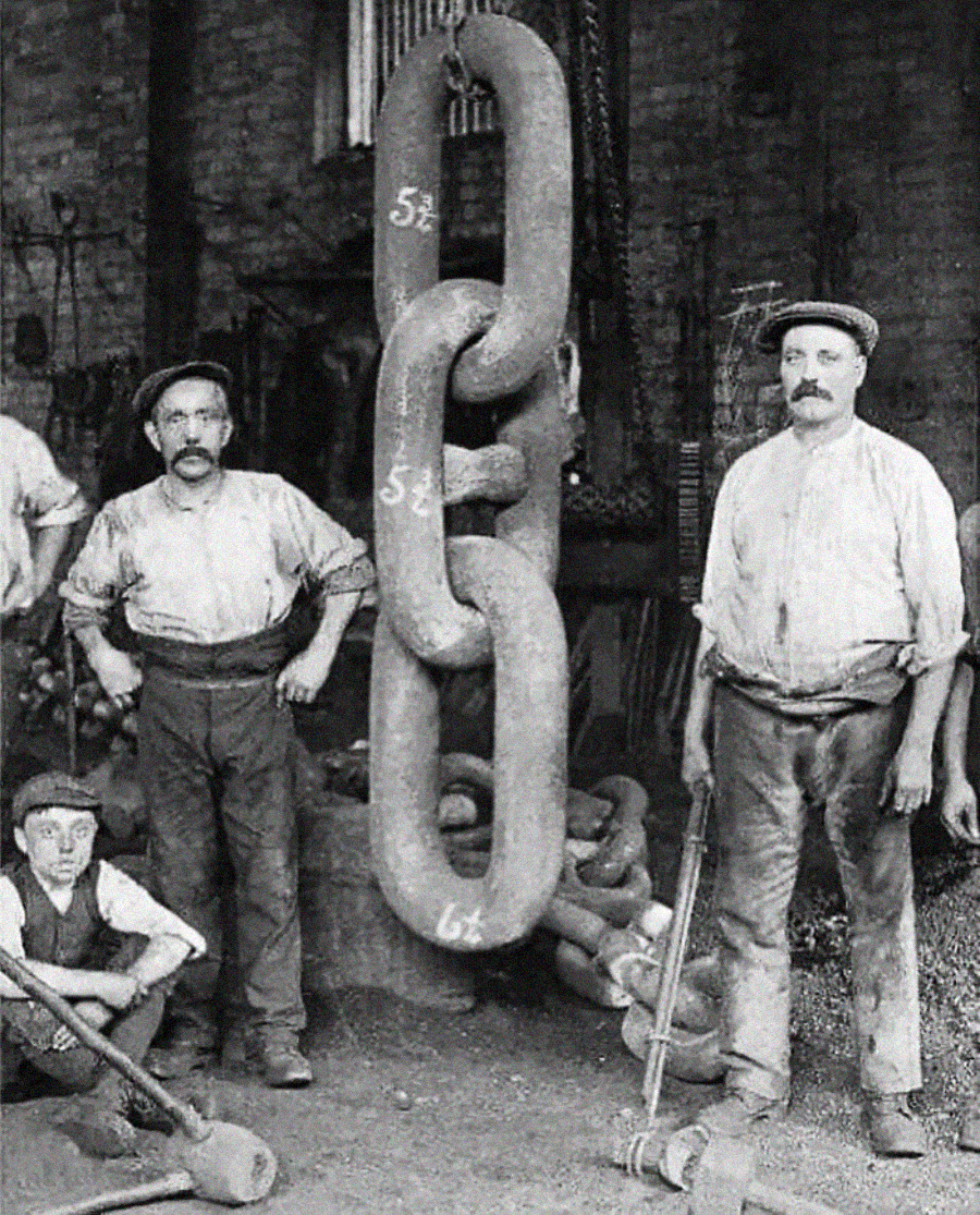 Four men in work clothes and caps stand around giant metal chain links in a workshop. Two men hold large hammers, and the oversized chain hangs vertically. The background shows industrial tools and a brick wall.